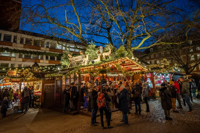 Ein beleuchteter Stand auf dem Weihanchtsmarkt in der Abenddämmerung. Davor stehen Besucherinnen und Besucher